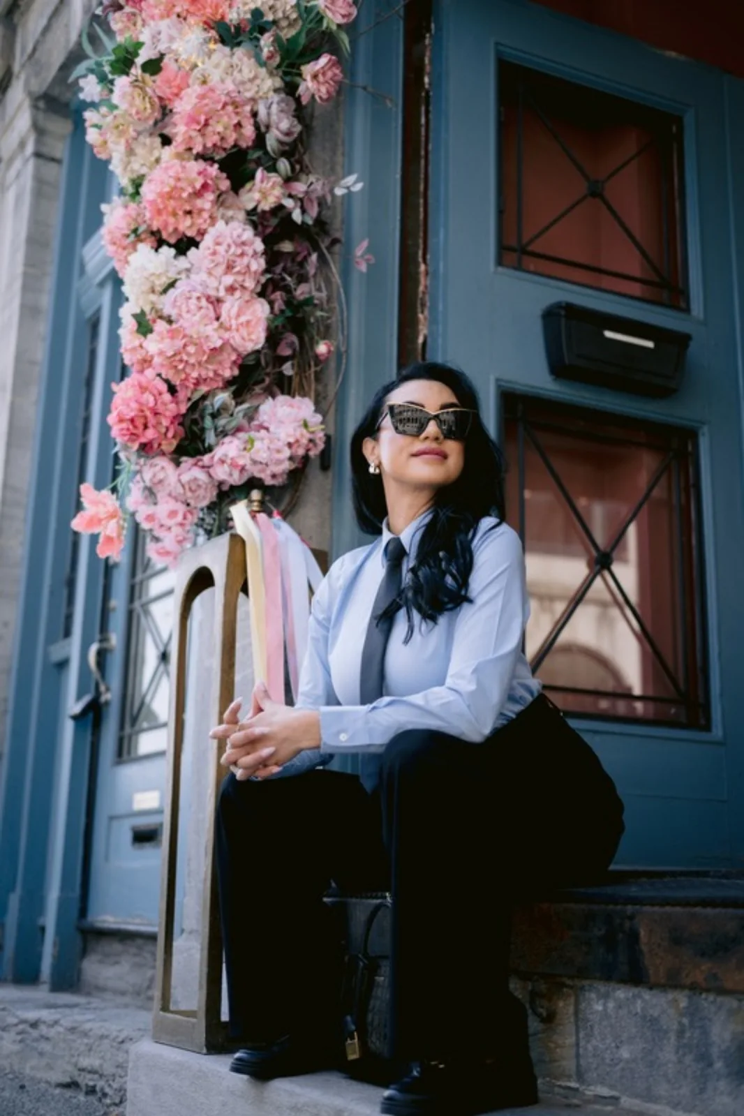 Stéphanie Nachar assise devant une porte bleue entourée de fleurs roses, en chemise et cravate.