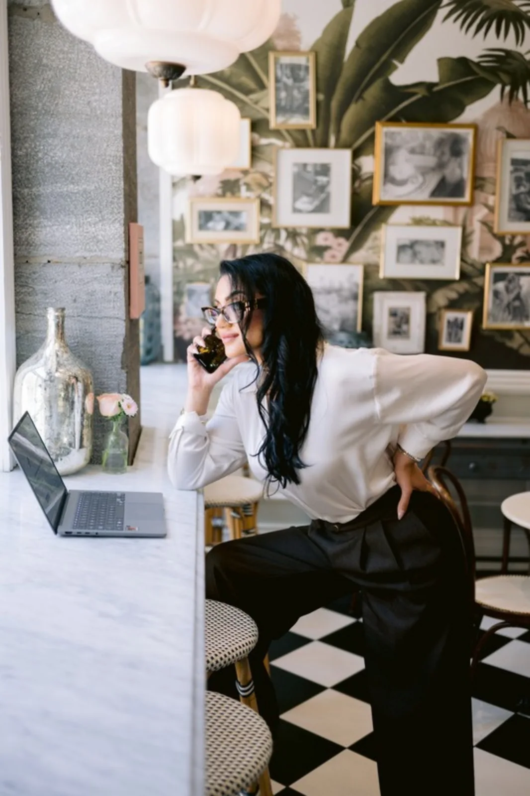 Stéphanie Nachar au téléphone dans un café élégant, devant un mur de photographies aux cadres dorés.